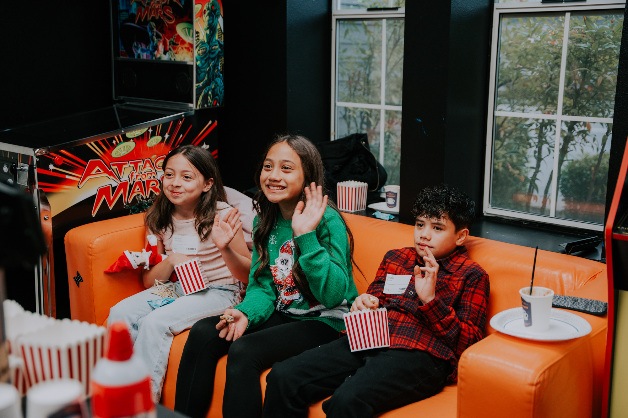Three children sitting on an orange couch waving and eating popcorn in a room with arcade games and large windows.