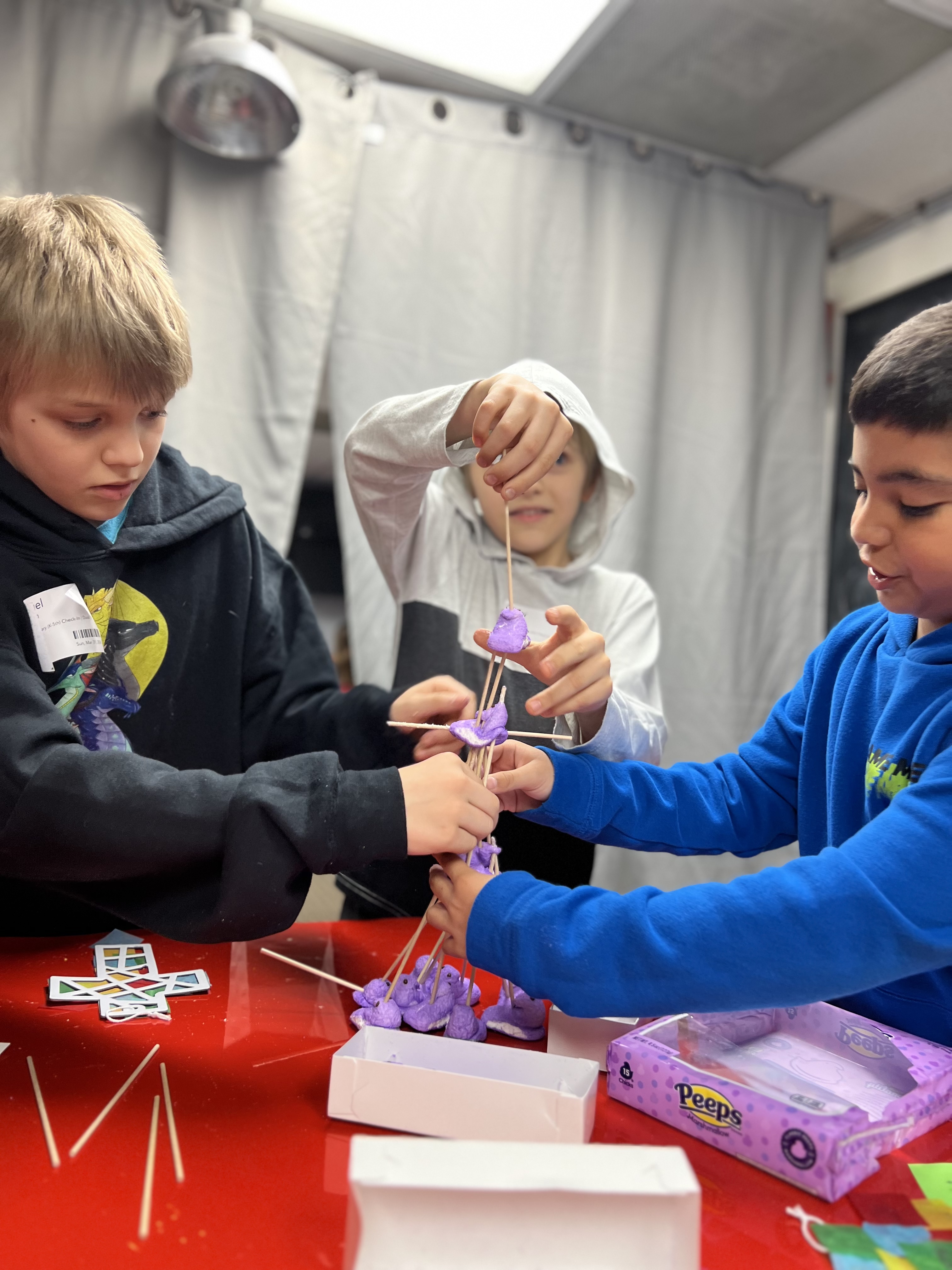 Three children collaboratively building a tall structure with purple marshmallow Peeps and wooden sticks on a red table.