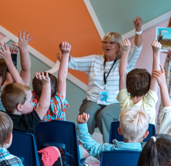 A group of children sitting in chairs raising their hands while an adult woman leads an activity in a colorful classroom.