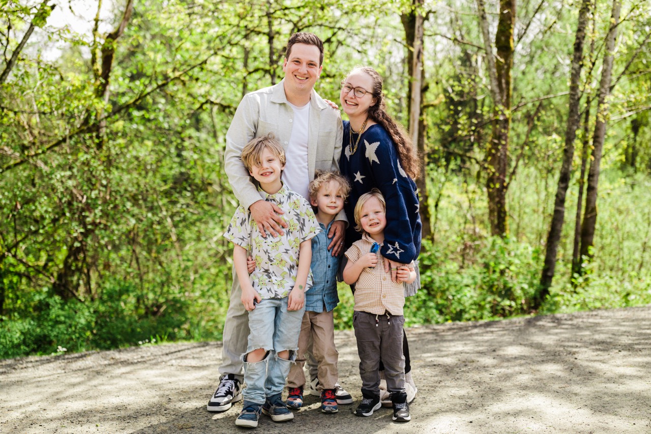 Smiling family of two adults and three young boys sitting on a blanket in a garden, with the boys wearing matching t-shirts.