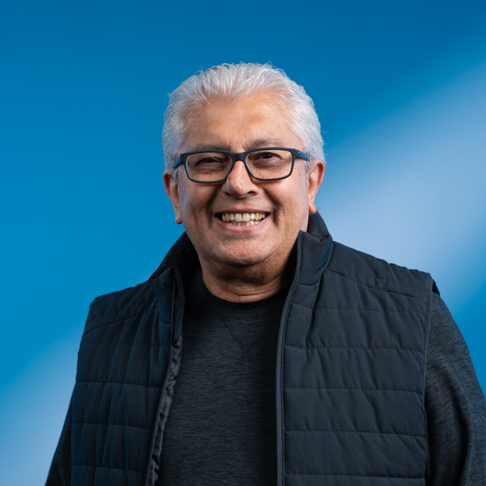 An elderly man with white hair and black rimmed glasses with brown eyes.