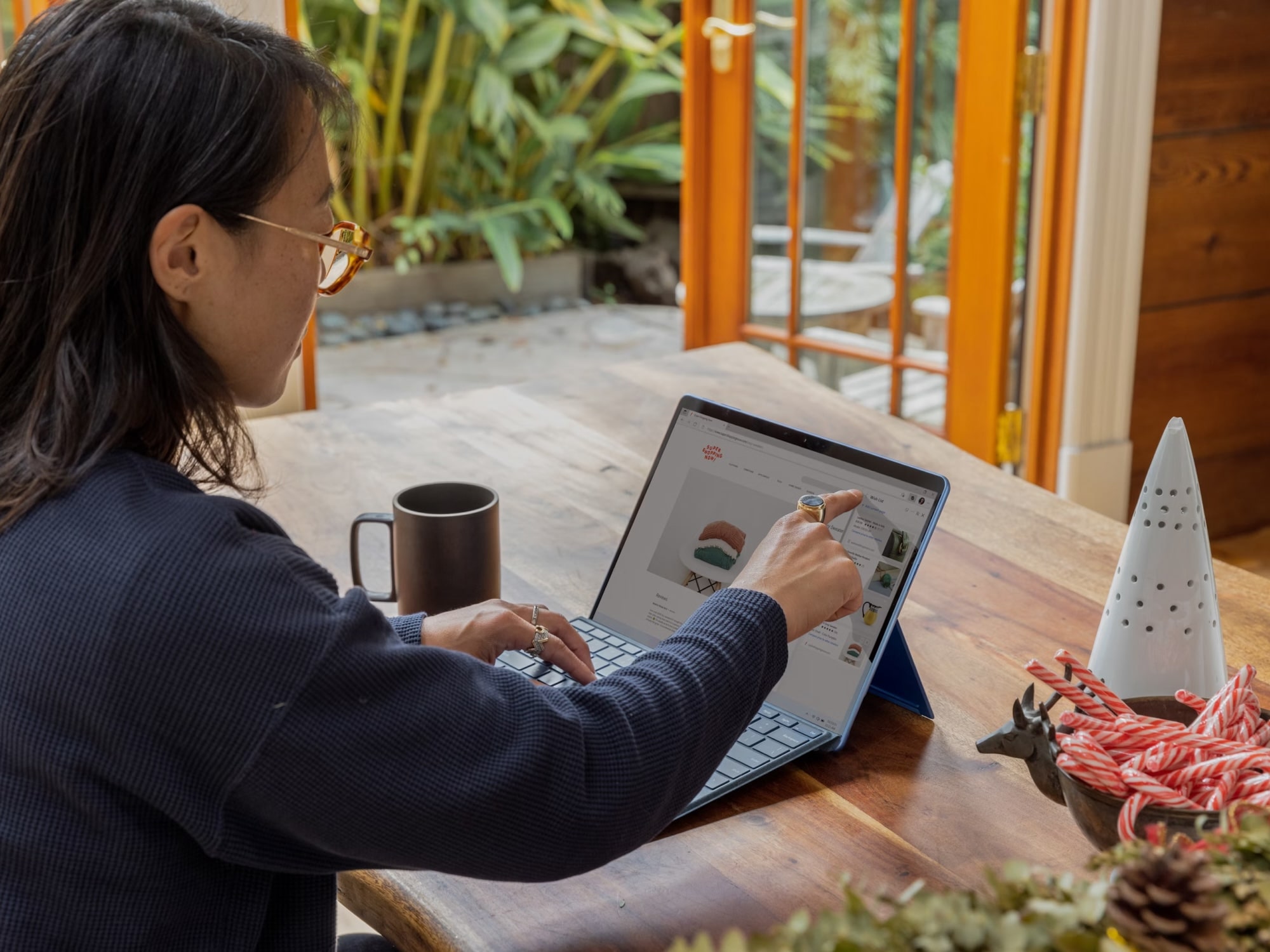 Person wearing glasses and a dark sweater interacting with a tablet device on a wooden table near a bowl of candy canes and festive decor.