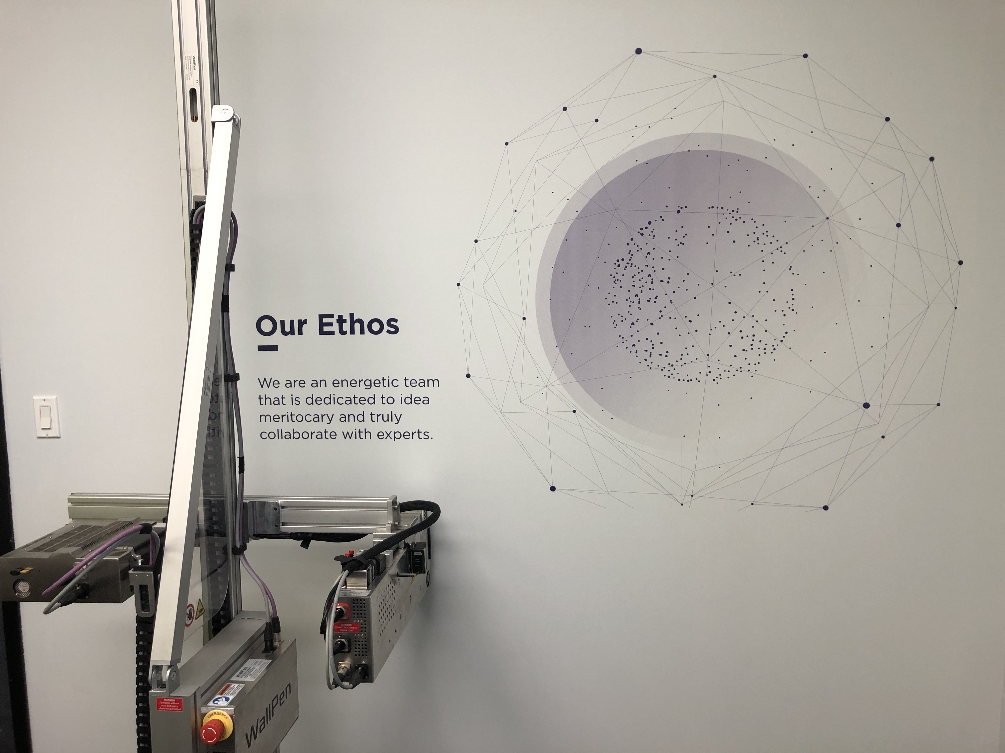 Wall with text 'Our Ethos' and a message about teamwork next to a geometric design with dots and lines, with a WallPen robotic device in the foreground.