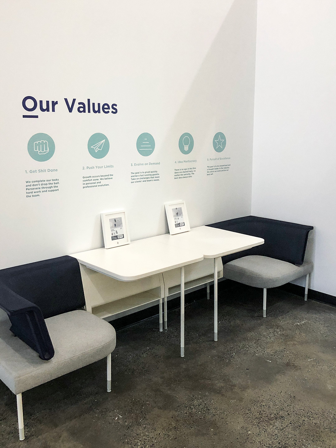 Two cushioned chairs facing white tables under a wall displaying five company values with icons, including 'Get Shit Done' and 'Push Your Limits'.
