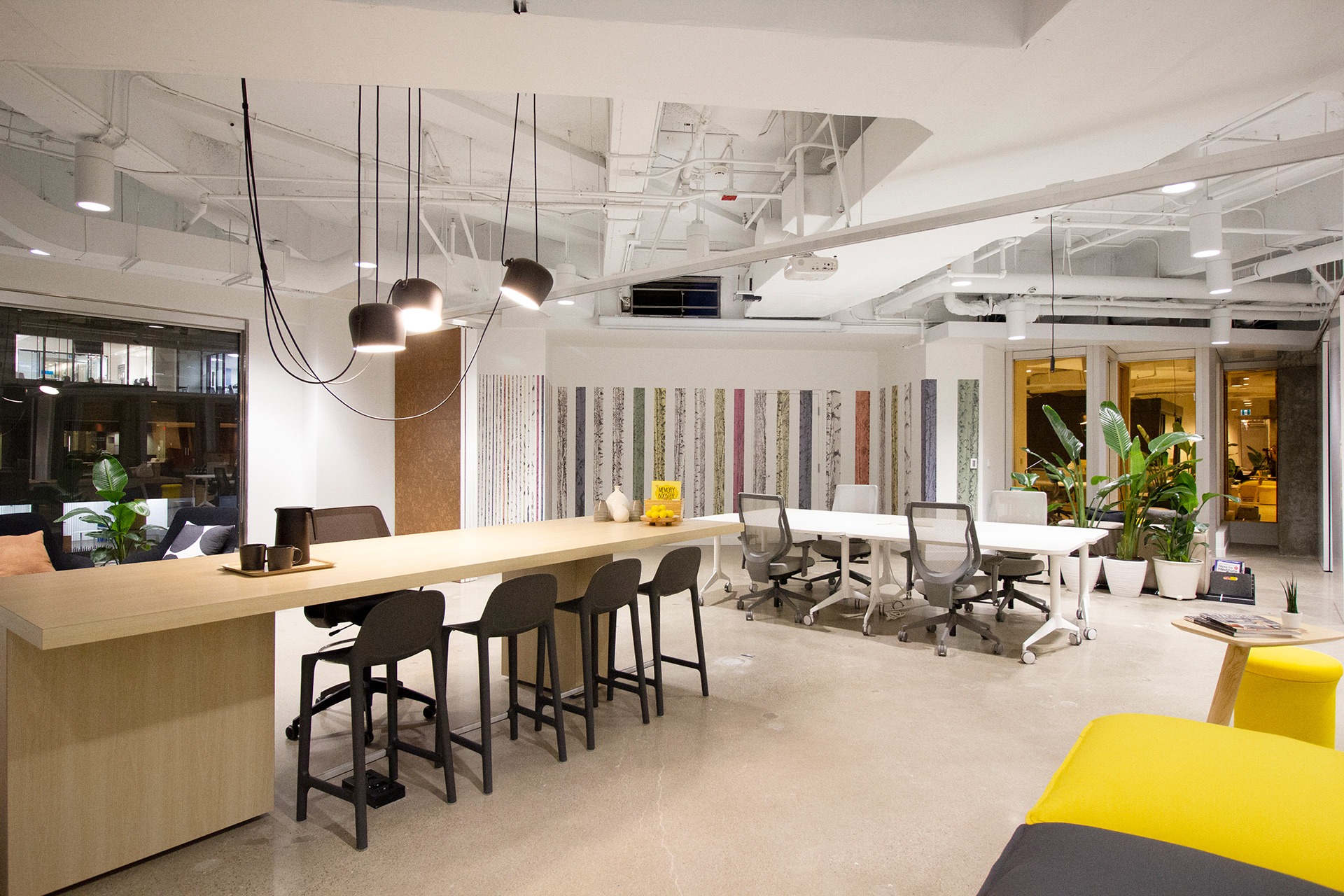 Modern open office space with wooden high table and black chairs, white work tables with gray office chairs, hanging lights, and potted plants.