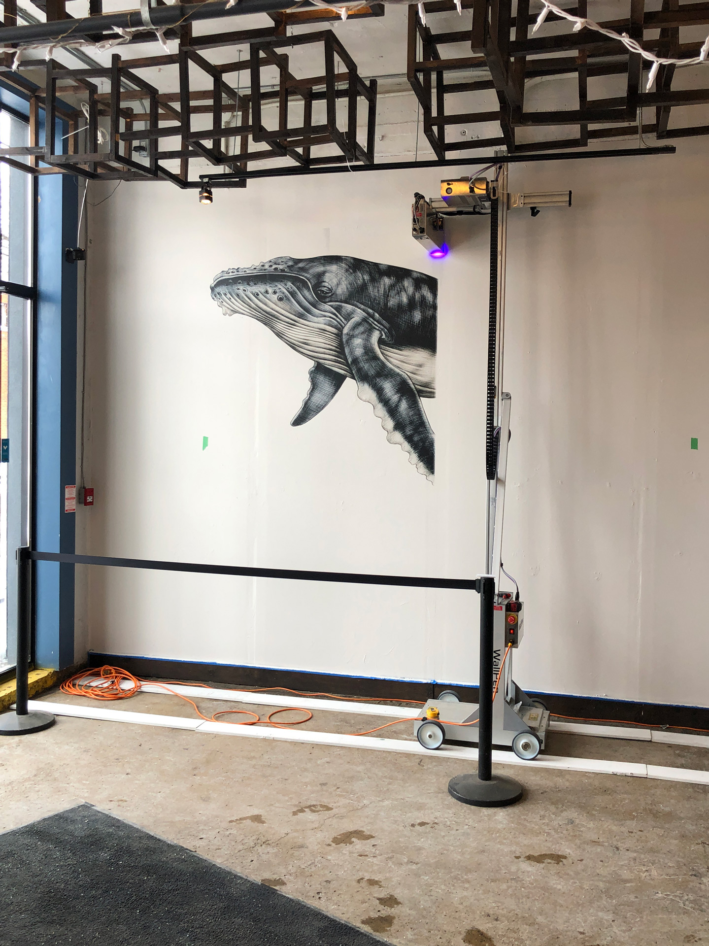 Black and white mural of a whale head and flipper on a white wall in an industrial-style room with metal ceiling frames and a robotic painting machine.