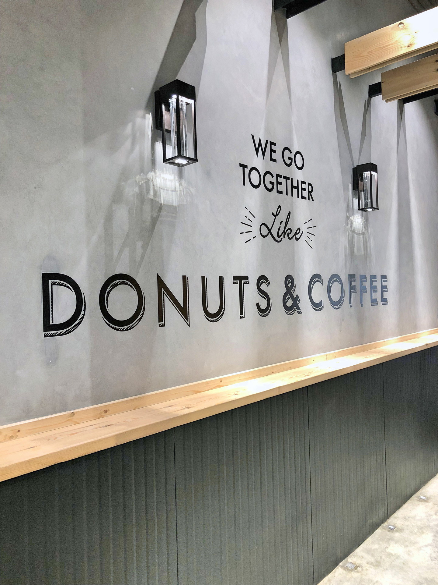 Interior wall with black text reading 'We go together like donuts & coffee' illuminated by two black wall lamps above a wooden ledge.