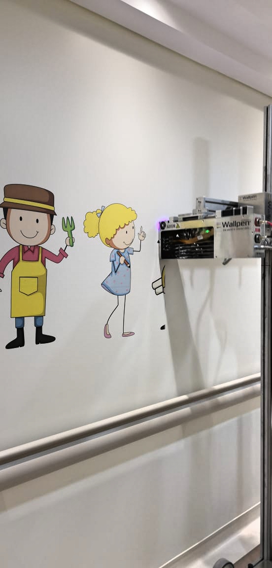 Wall painting of cartoon characters, including a boy in a yellow apron holding a garden fork and a girl with blonde hair in a blue dress, with a Wallpen device mounted on the wall printing part of the image.