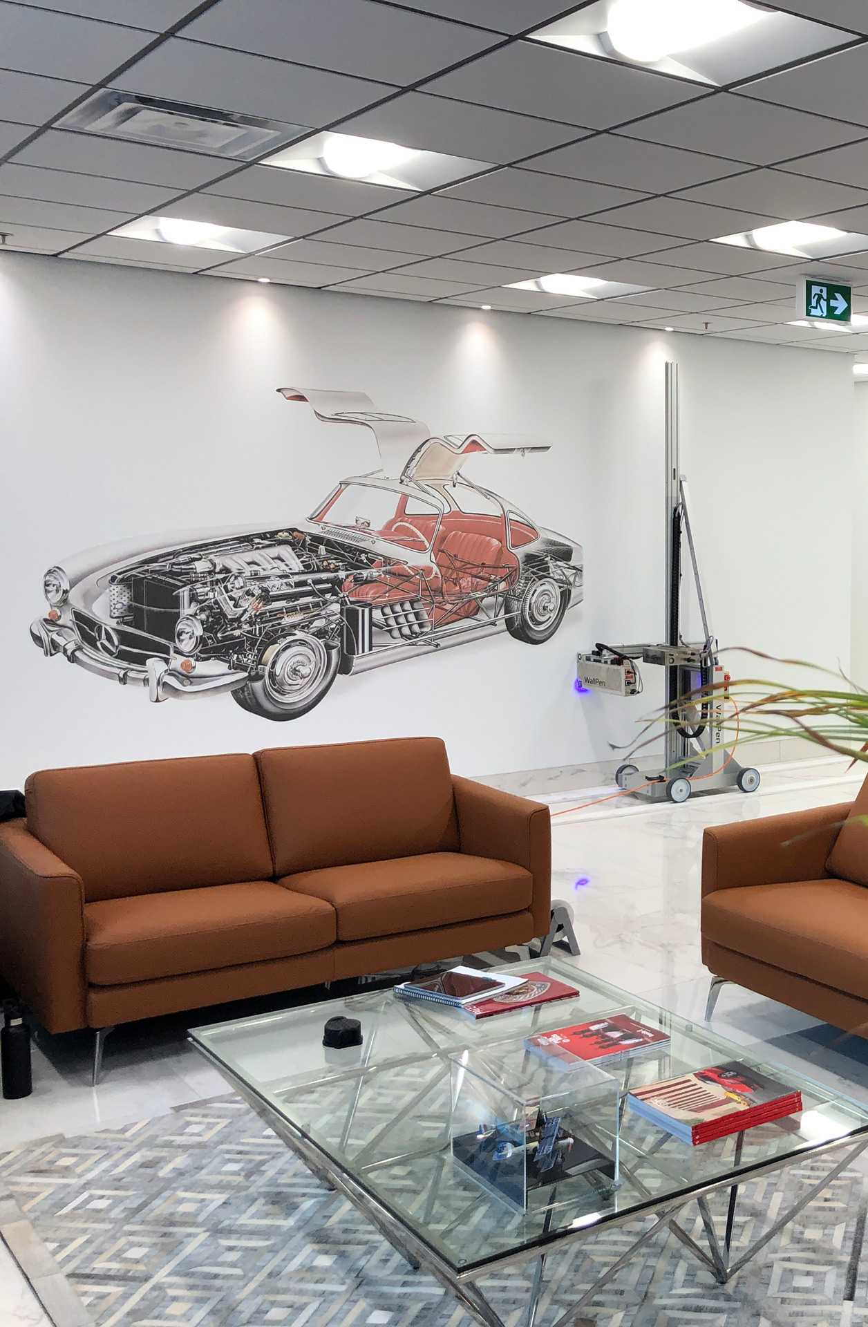 Modern office lounge with brown sofas, glass coffee table with books, and a wall mural of a vintage Mercedes car showing internal components.