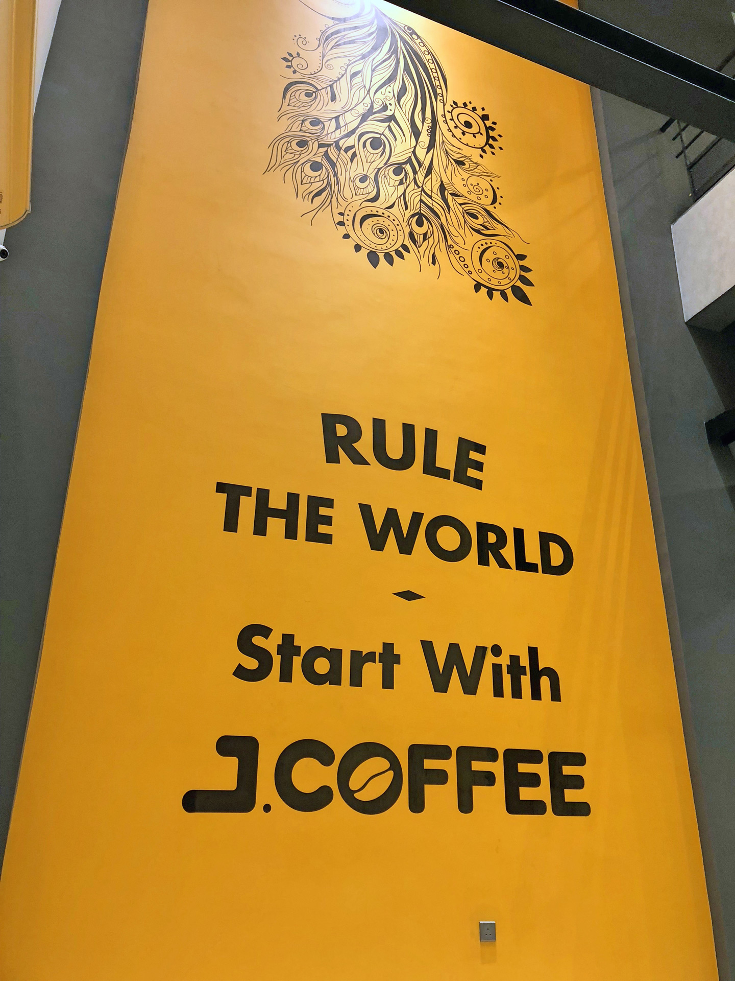 Large yellow wall banner with black text saying 'RULE THE WORLD - Start With J.COFFEE' and a decorative peacock feather design at the top.