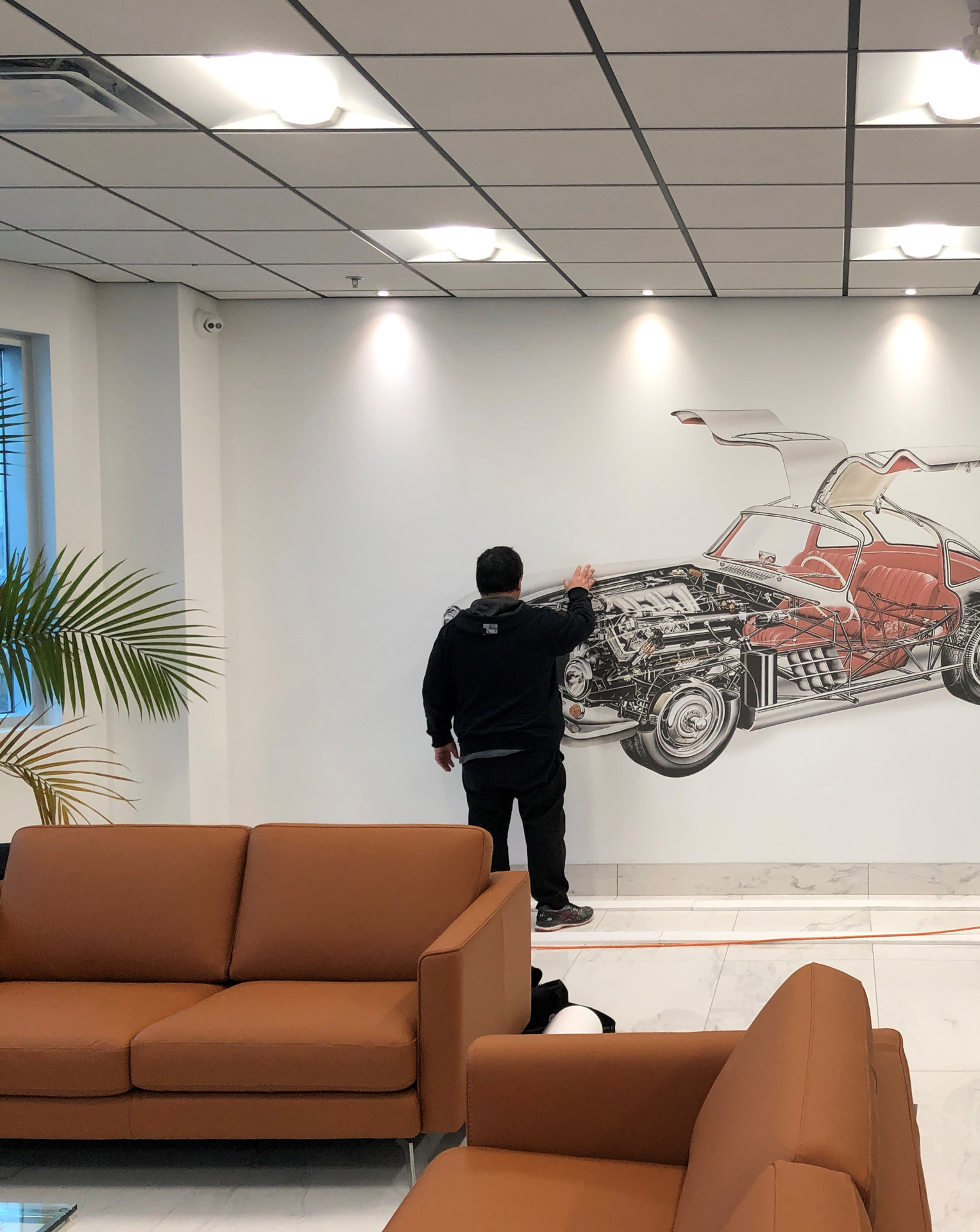 Person in black clothing touching a detailed wall mural of a car with gull-wing doors in a modern room with brown sofas and a potted plant.