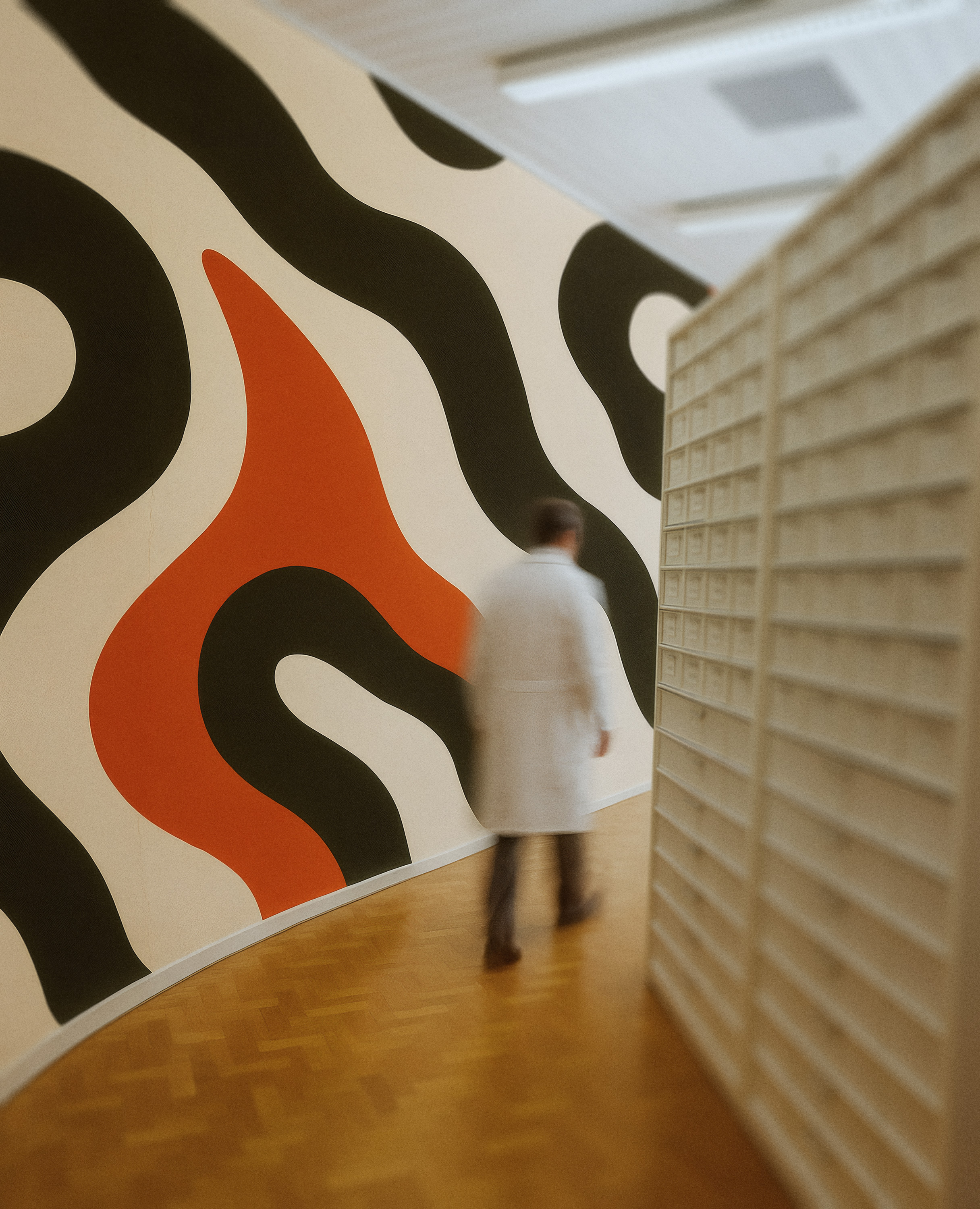 Blurry figure of a person in a white coat walking along a wooden floor corridor with a colorful abstract mural on the wall.