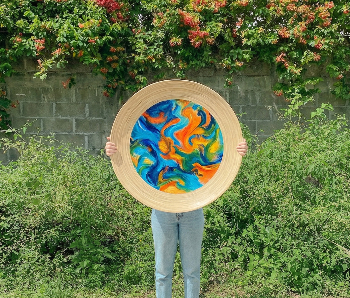 Person holding a large round frame with a colorful swirling abstract painting in front of their torso, standing outdoors in front of green foliage and a brick wall.