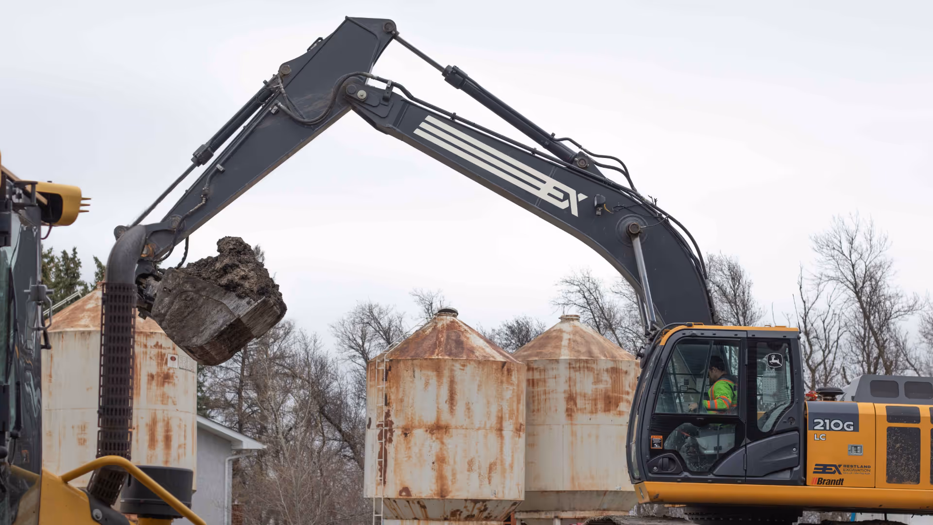 Excavator lifting a bucket full of dirt at a construction site with rusted storage tanks and leafless trees in the background.