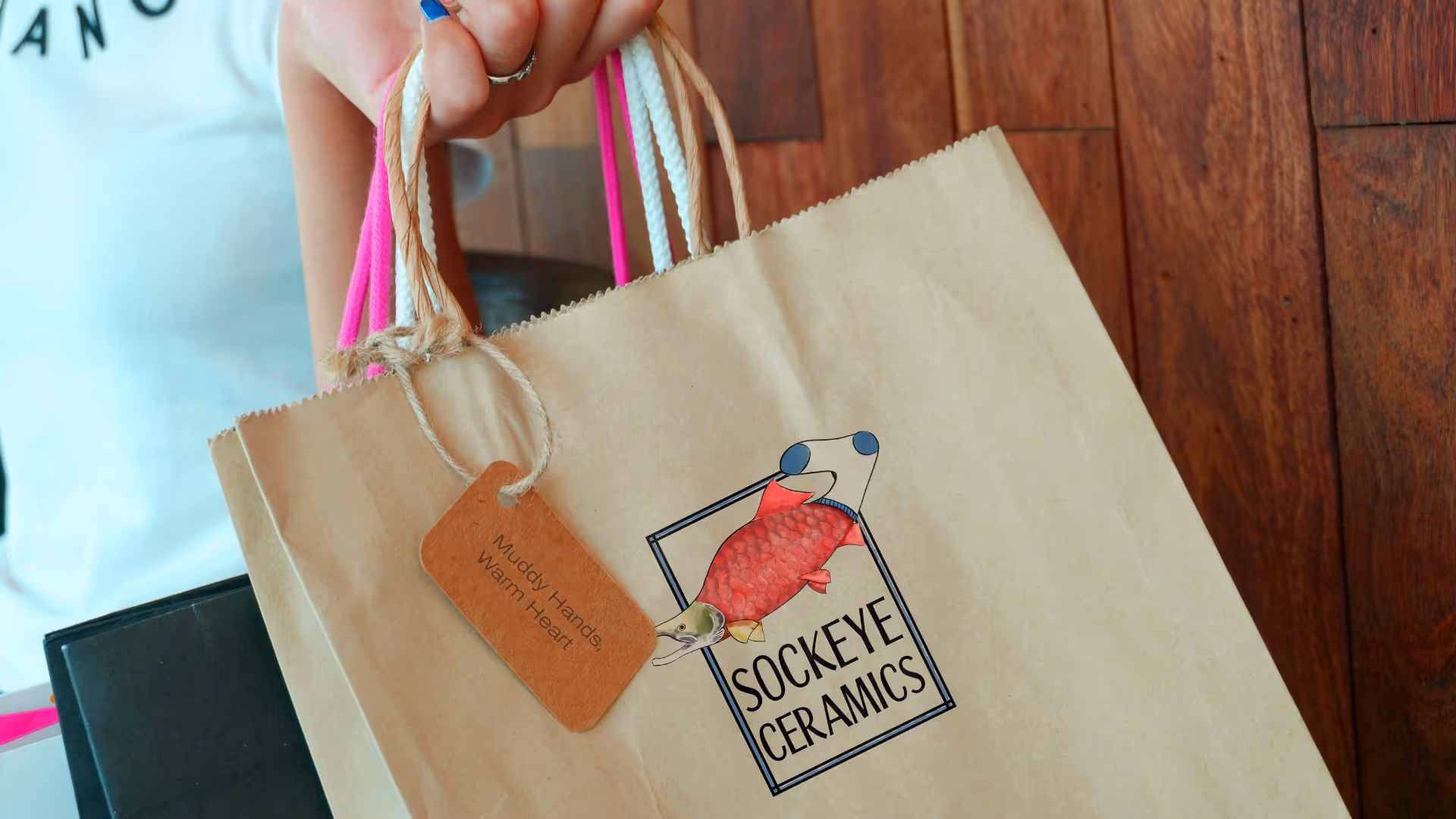 Hand holding brown paper shopping bag with a colorful fish logo and tag reading 'Muddy Hands, Warm Heart'.