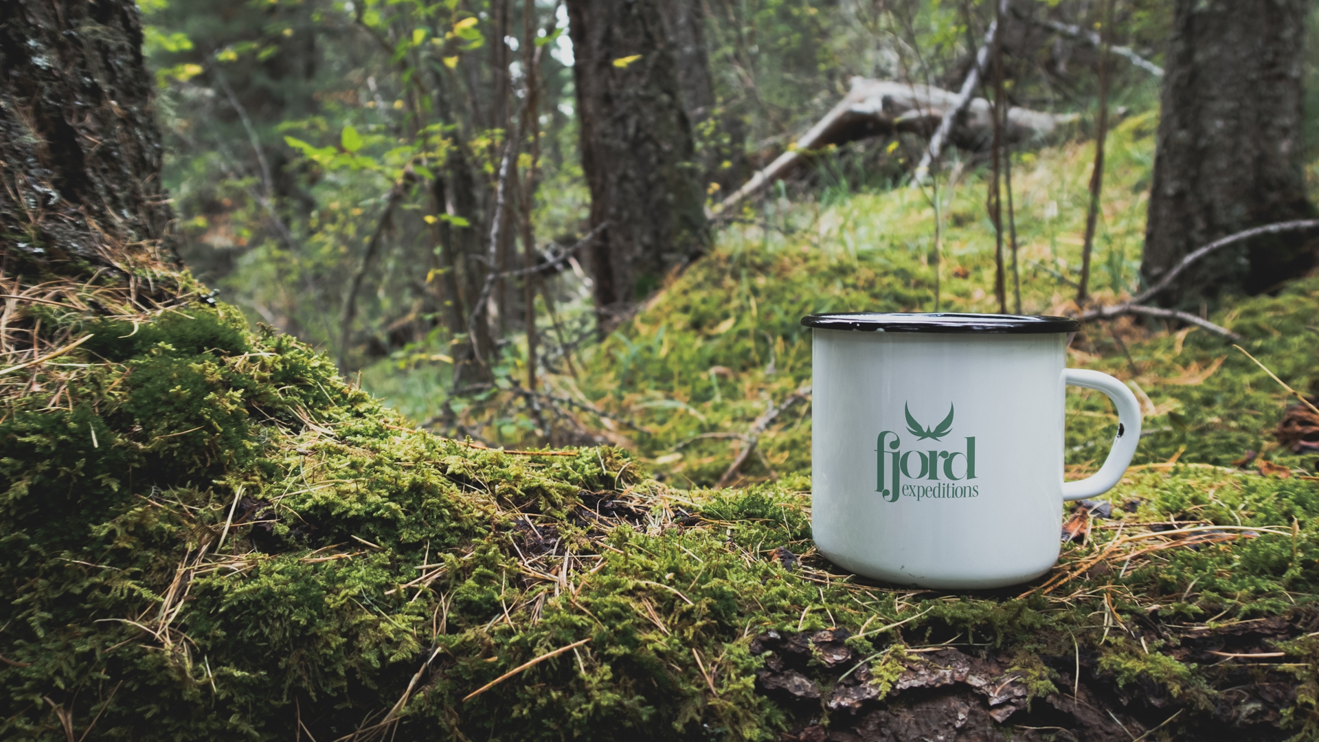 White metal mug with Fjord Expeditions logo placed on moss-covered forest floor.