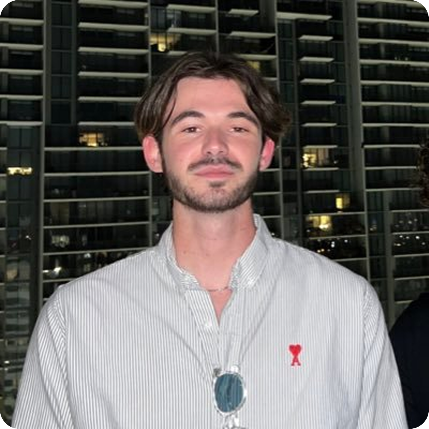 Young man with short brown hair and beard wearing a light striped shirt with a red emblem, standing in front of a lit building at night.
