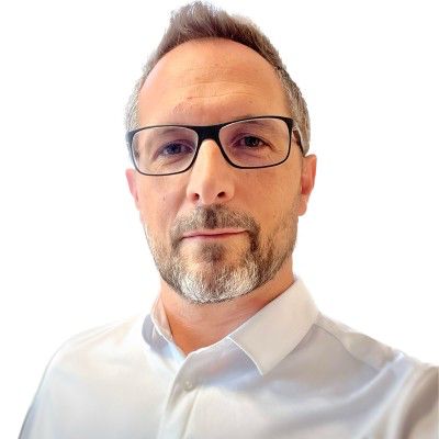 Middle-aged man with glasses and short graying beard wearing a white collared shirt against a white background.