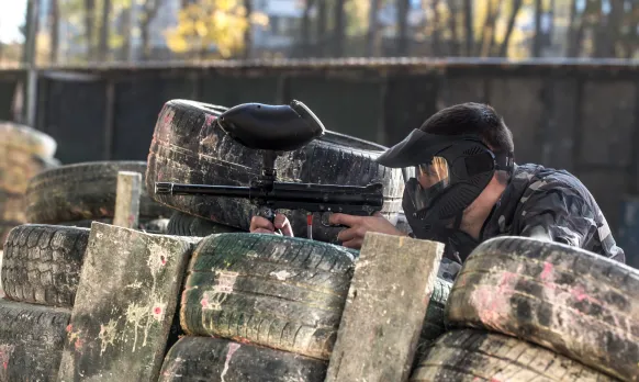 Person wearing a protective mask aiming a paintball gun behind stacked tires at a paintball field.