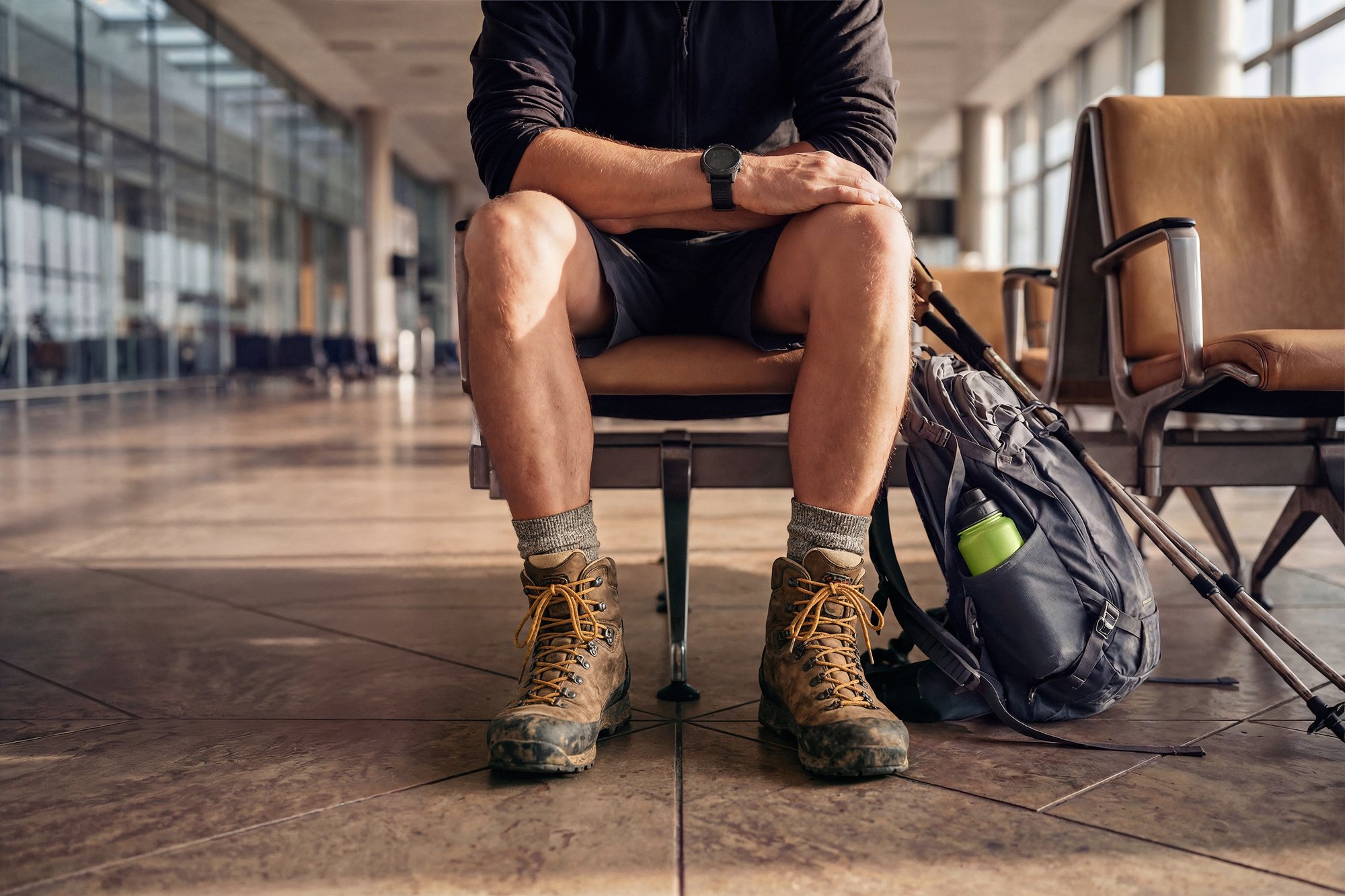 The Trail: a traveller in worn hiking boots and shorts waiting at an airport gate beside a backpack and trekking poles, shown from the knees down - AI-generated image by Matter Studio for Zurich Travel Guard