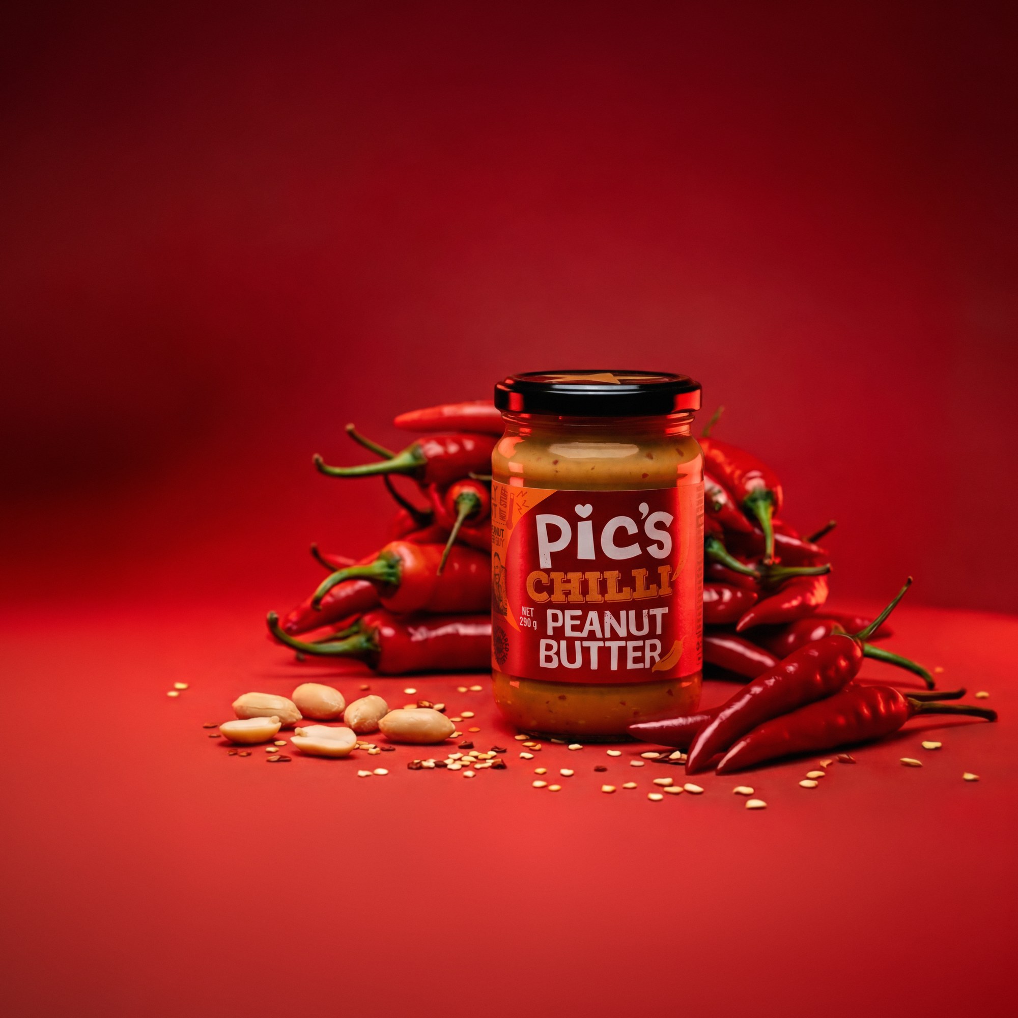 Pic's Chilli Peanut Butter jar in front of a pile of fresh red chillies with peanuts and chilli flakes on a deep red background - product image built by Matter Studio from a pre-production jar