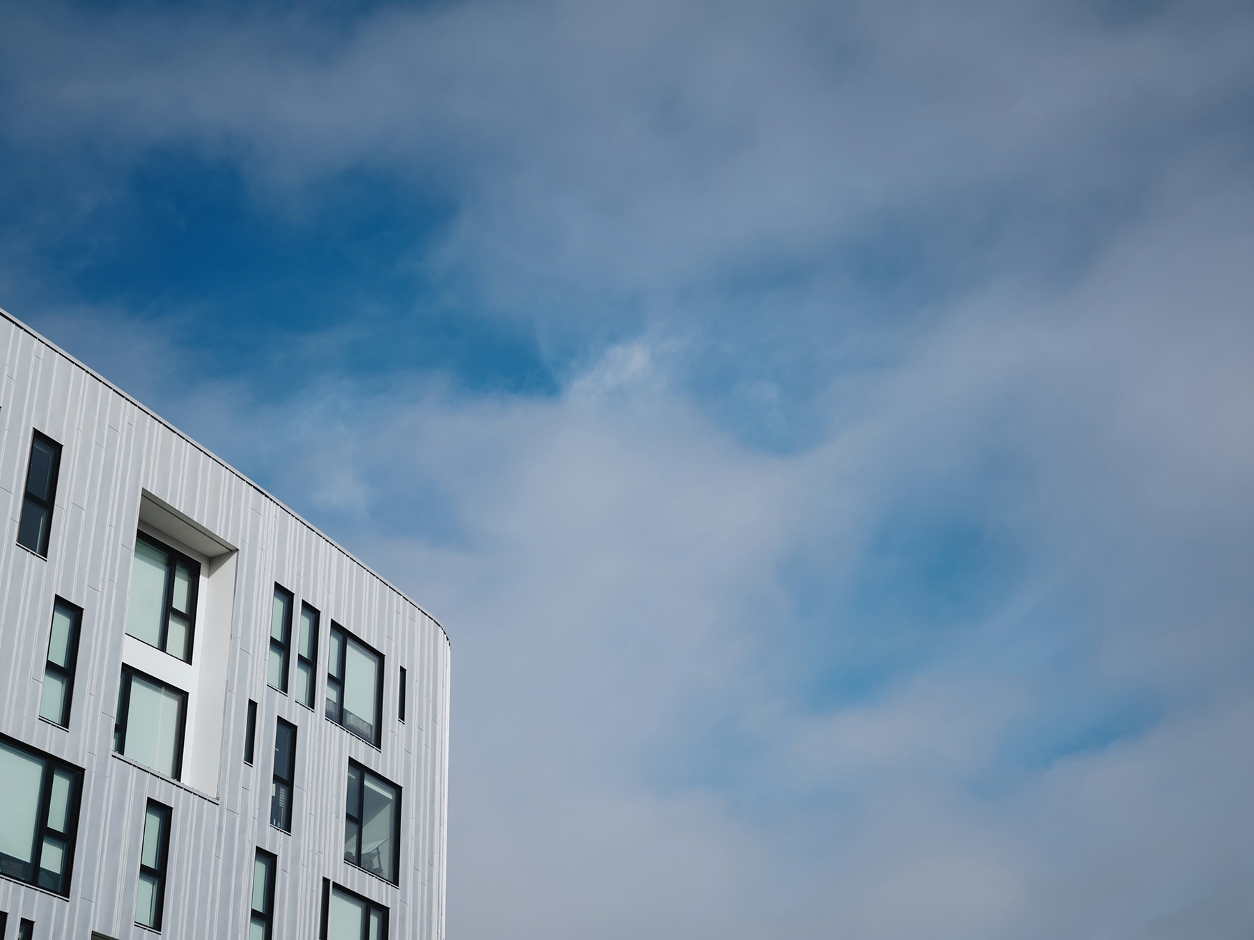 Modern white building facade with variously sized windows against a partly cloudy blue sky.