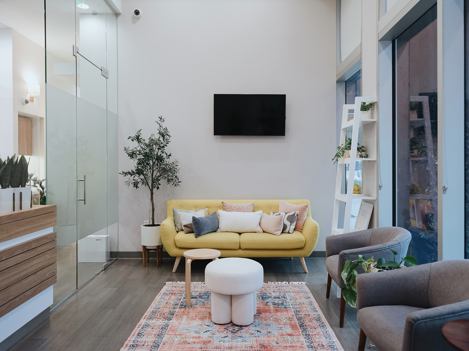 Modern living room with a yellow sofa, assorted pillows, two gray armchairs, a round white ottoman, wooden side table, plants, a patterned rug, and a wall-mounted flat-screen TV.