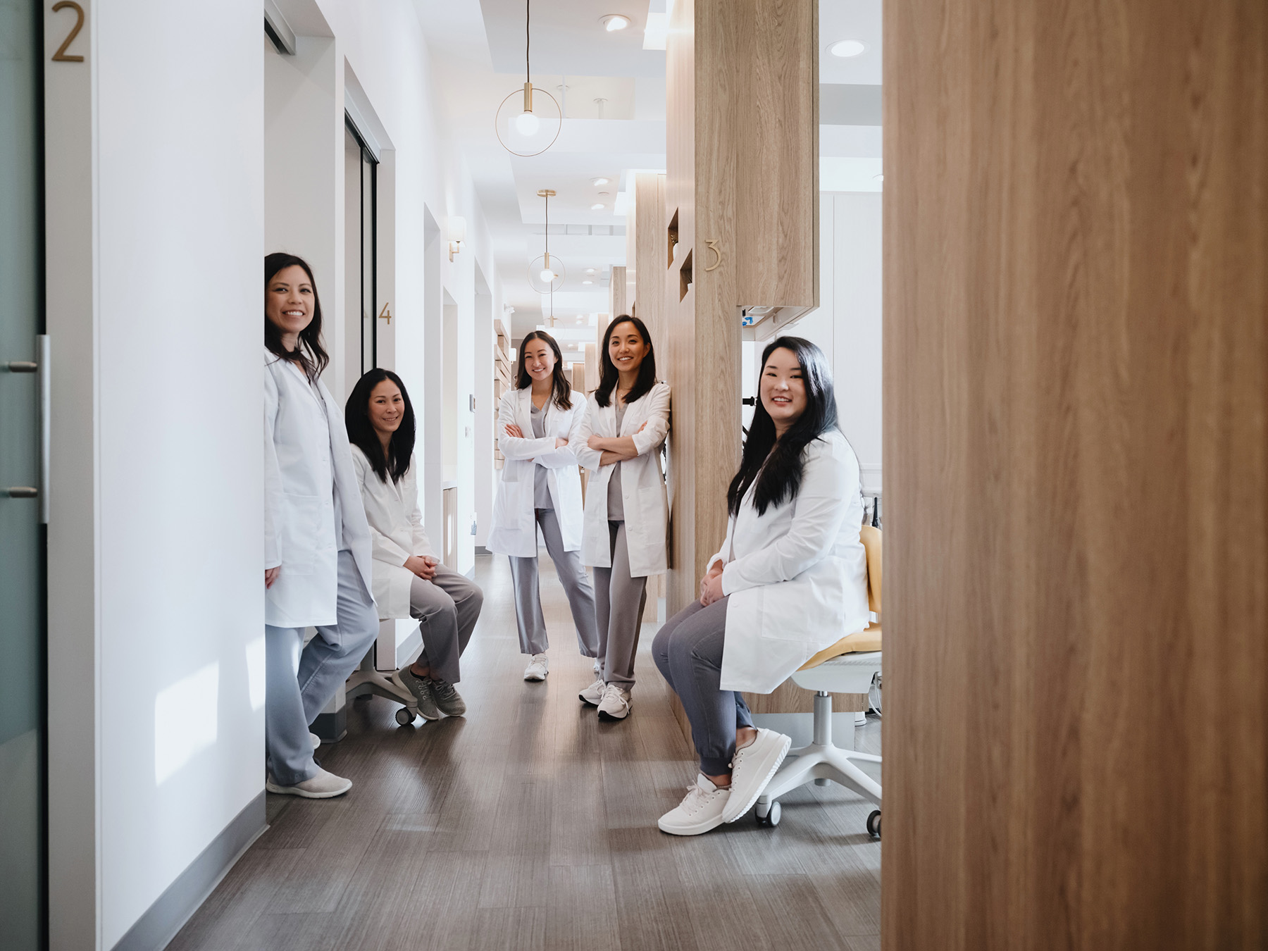 Five female medical professionals in white coats smiling and posing in a modern clinic corridor.
