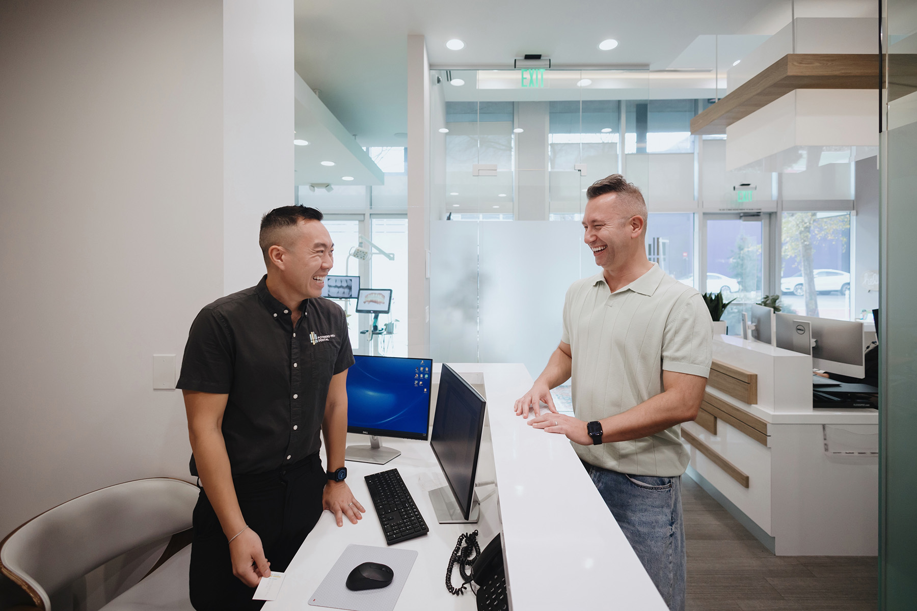 Two men smiling and laughing while talking across a reception desk in a modern office.