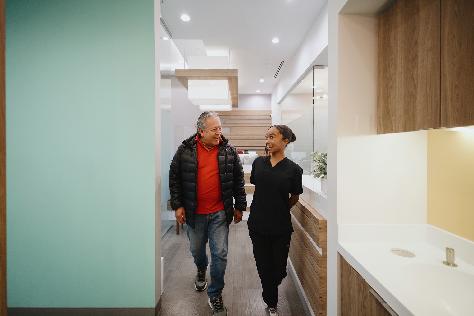 Man in a black jacket and red shirt walking and smiling with a woman in black scrubs in a modern medical office hallway.