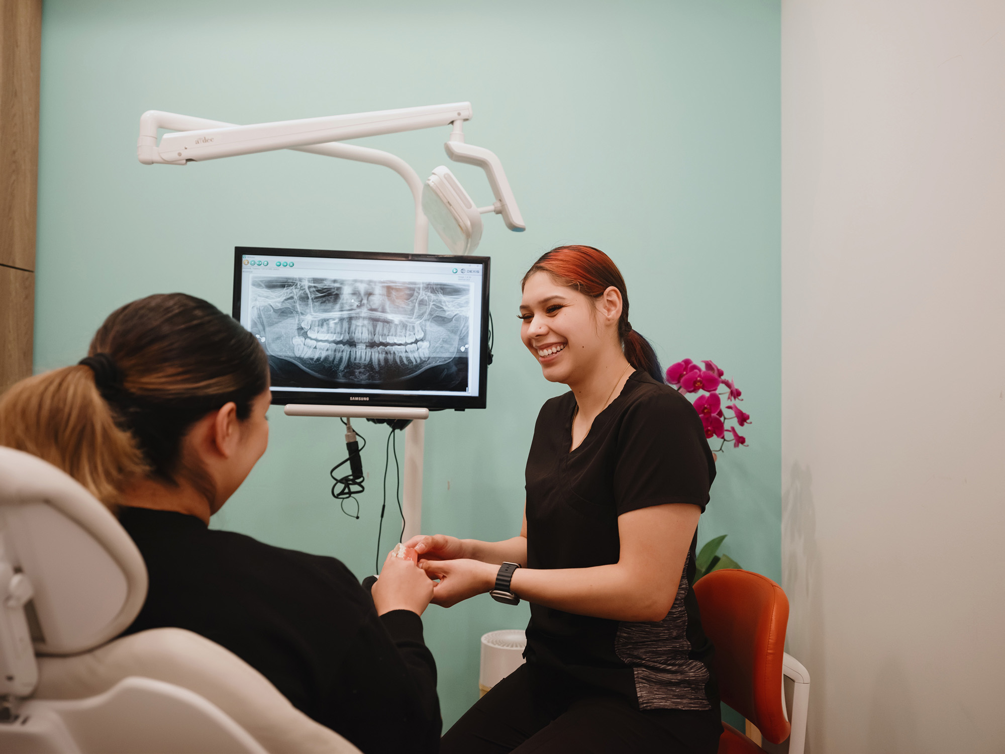 Dentist smiling and holding a dental model while consulting with a patient, with a dental X-ray displayed on a monitor in the background.