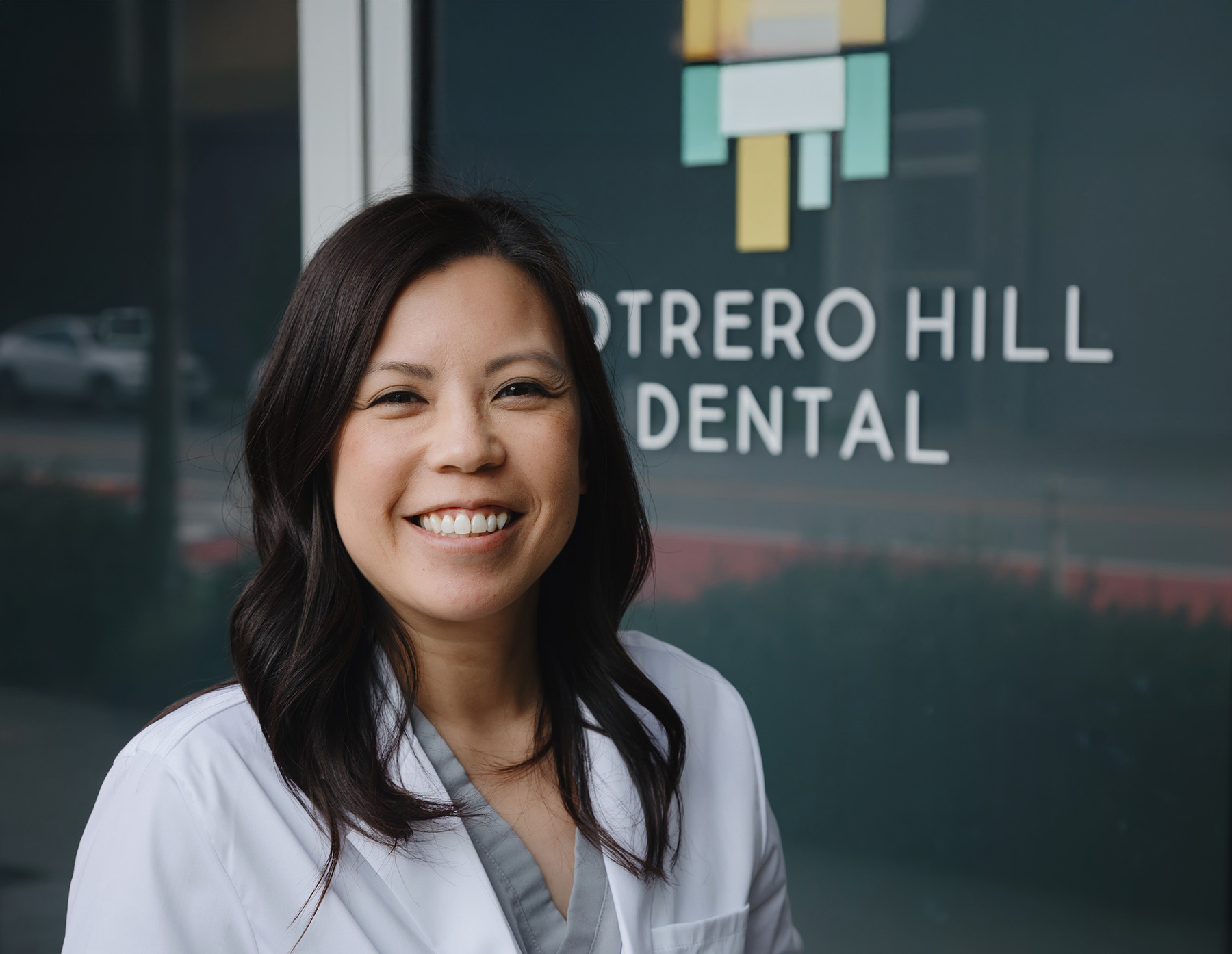 Smiling female dentist with dark hair in a white coat standing in front of a window with Potrero Hill Dental sign.