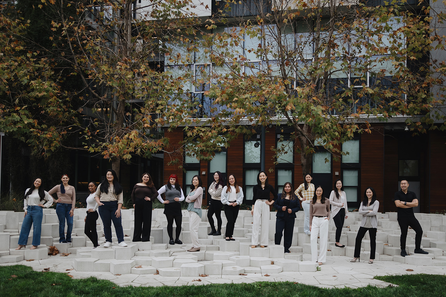 Group of diverse professionals posing outdoors on modern stone steps in front of glass-paneled building with autumn trees overhead.