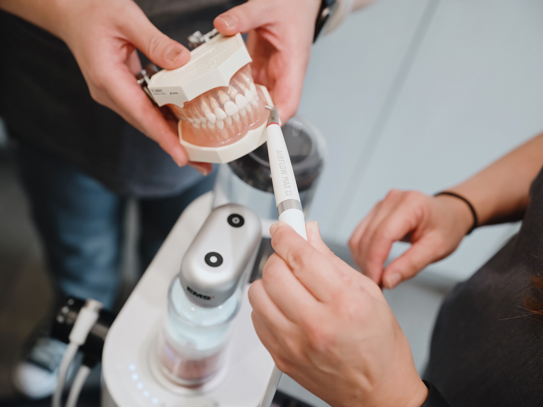 Hands holding dental model and Airflow Max device near dental equipment.