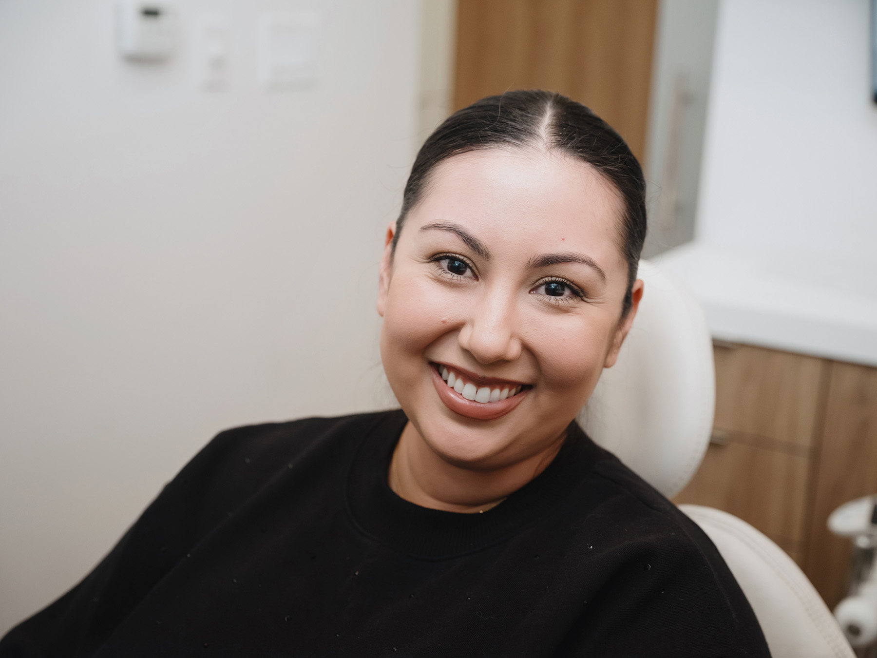 Smiling young woman with dark hair pulled back, wearing a black top, seated in a dentist's chair.