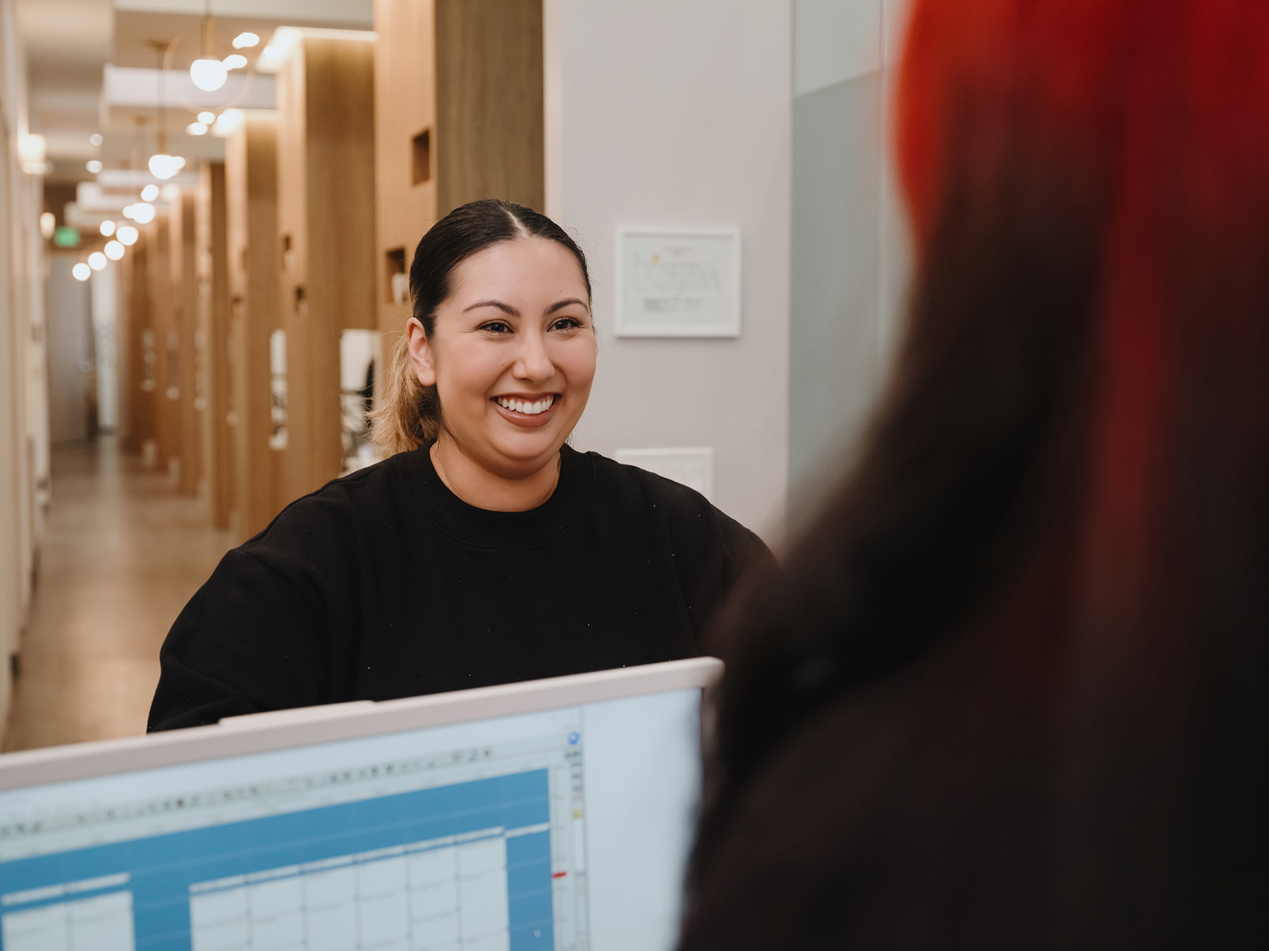 Smiling woman with hair tied back standing in an office corridor, facing a computer screen.