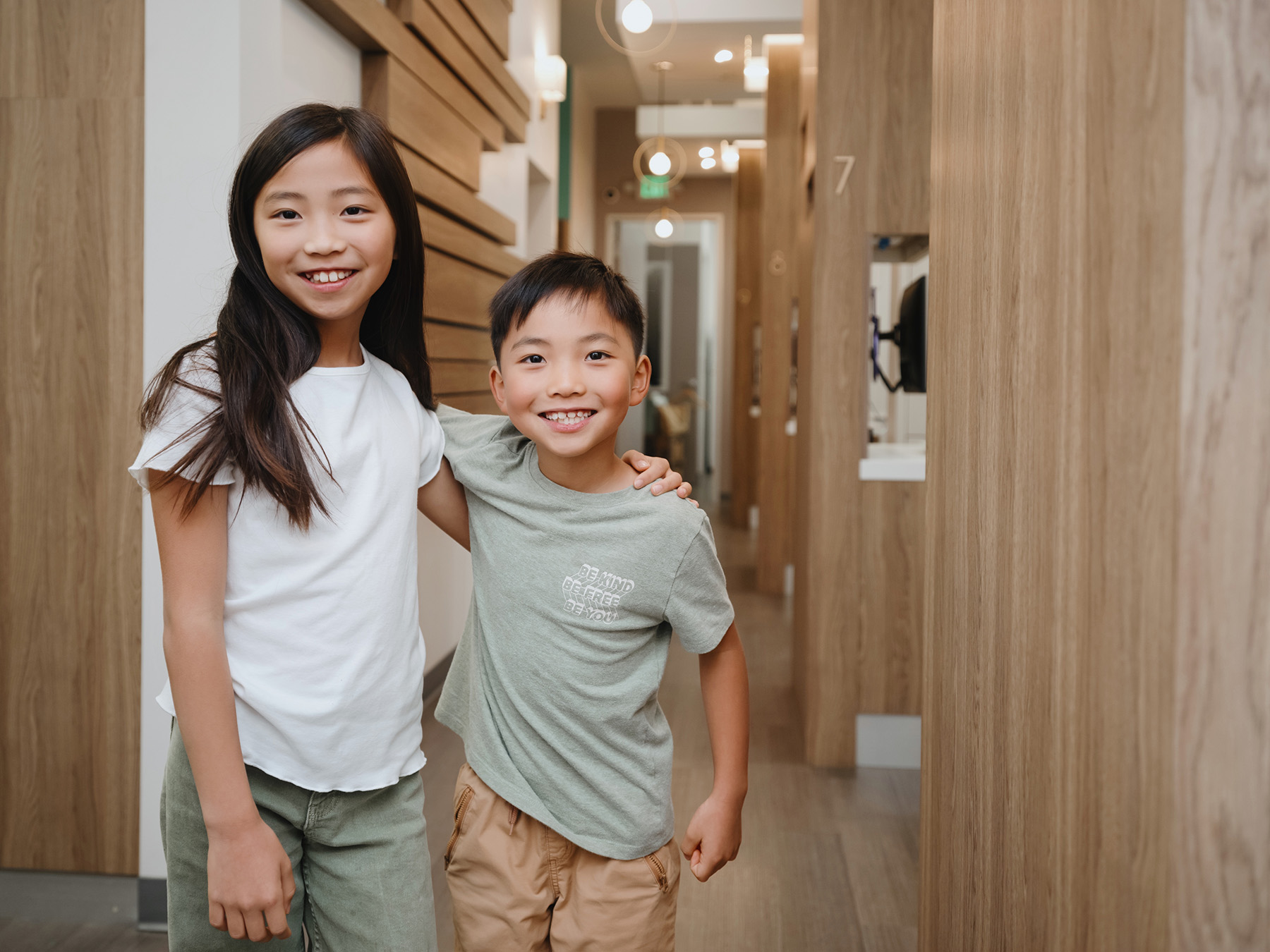 Smiling young girl with long dark hair and boy with short hair standing in a wood-paneled hallway, boy's arm around girl's shoulder.
