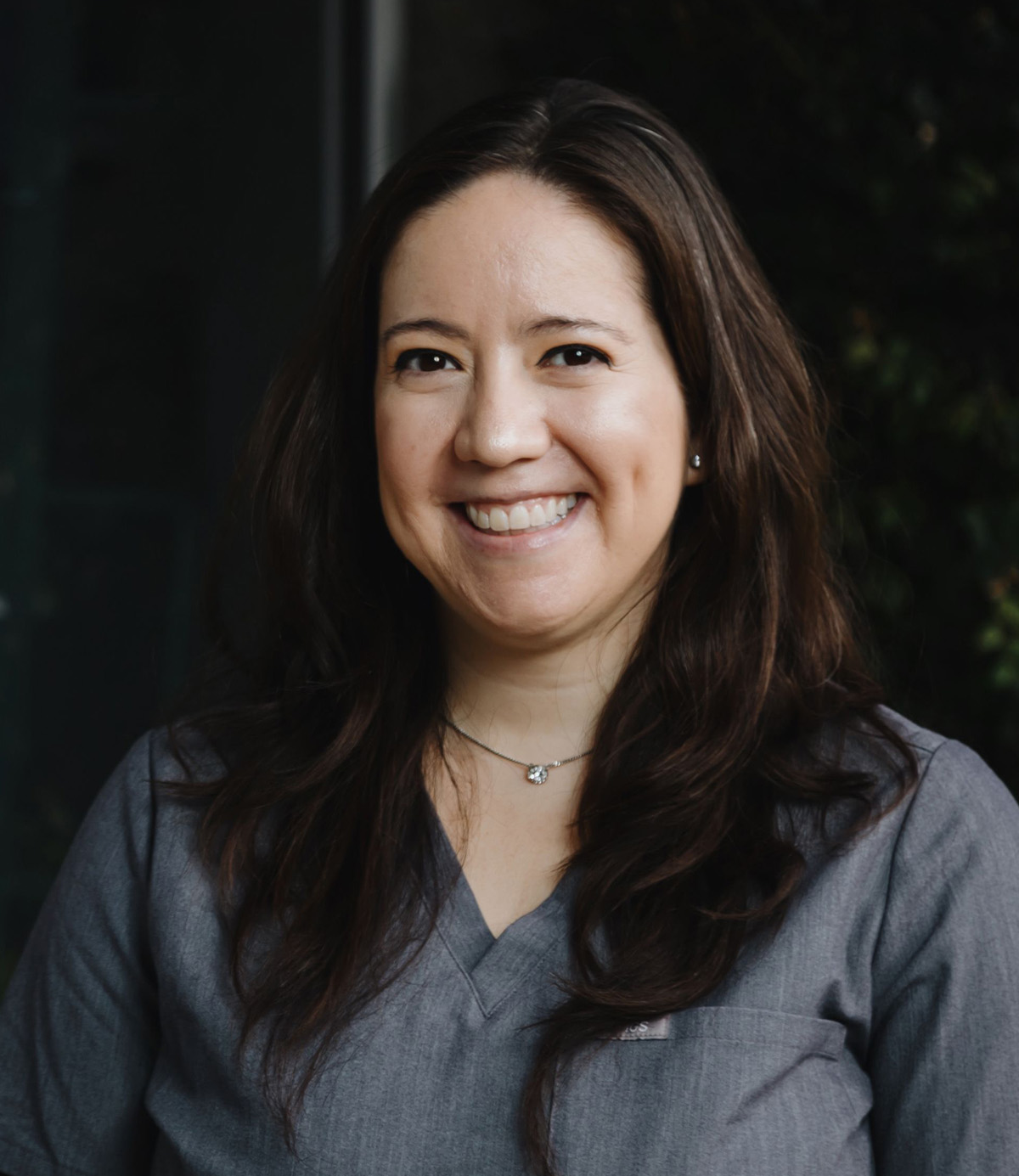 Smiling woman with long brown hair wearing a gray V-neck top and a necklace with a small pendant.