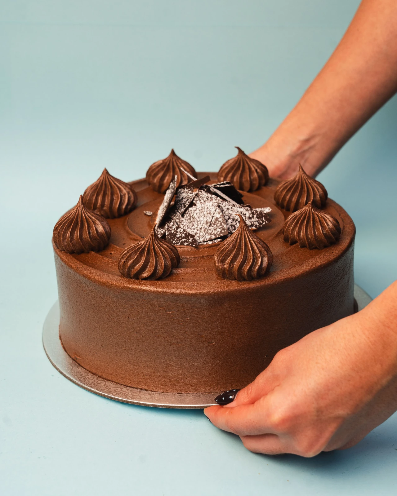 A hand setting down a chocolate layer cake on a pale blue studio backdrop