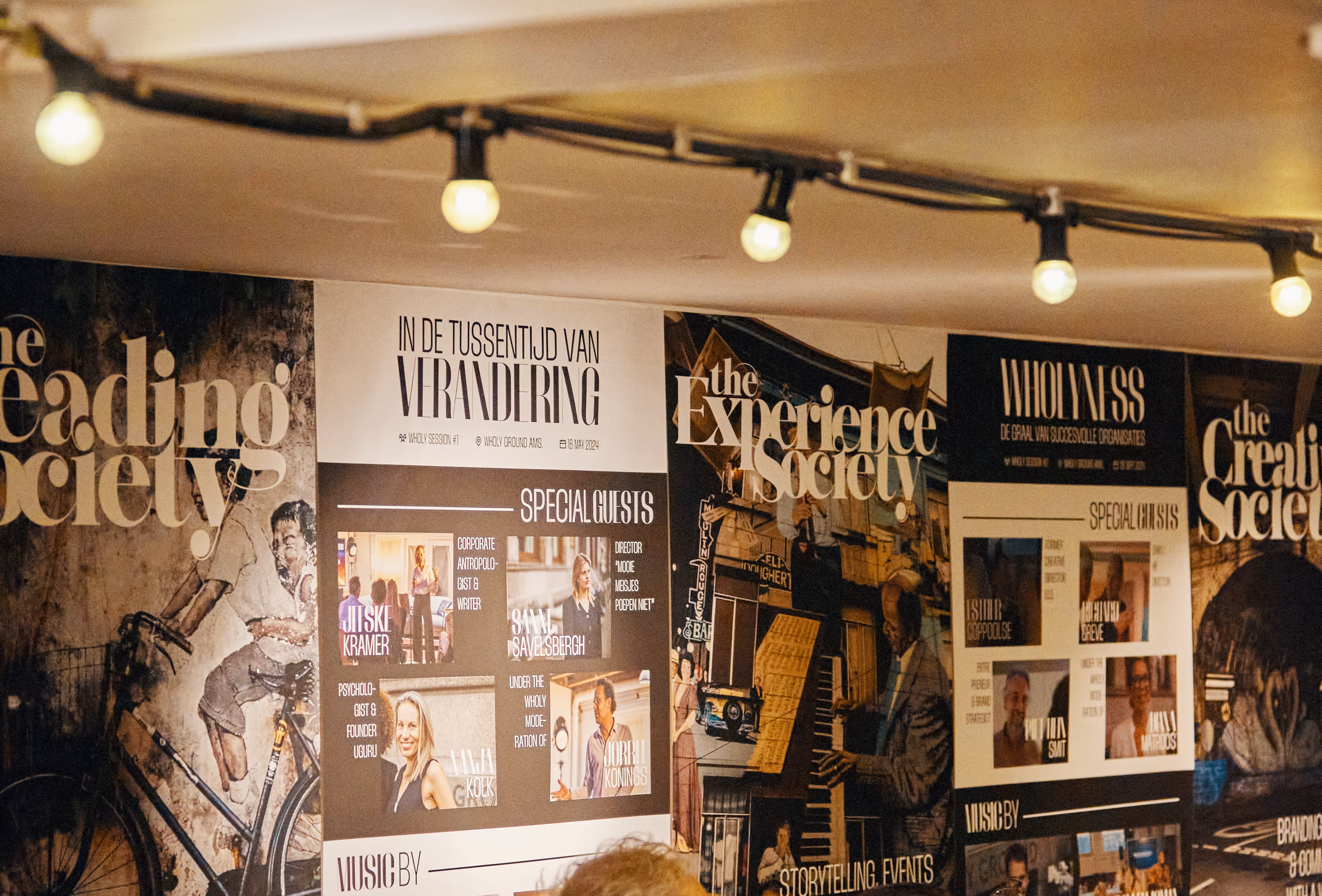 Wand met posters van evenementen en sessies met teksten over verandering, speciale gasten en creatieve samenlevingen, verlicht door een rij hanglampen.
