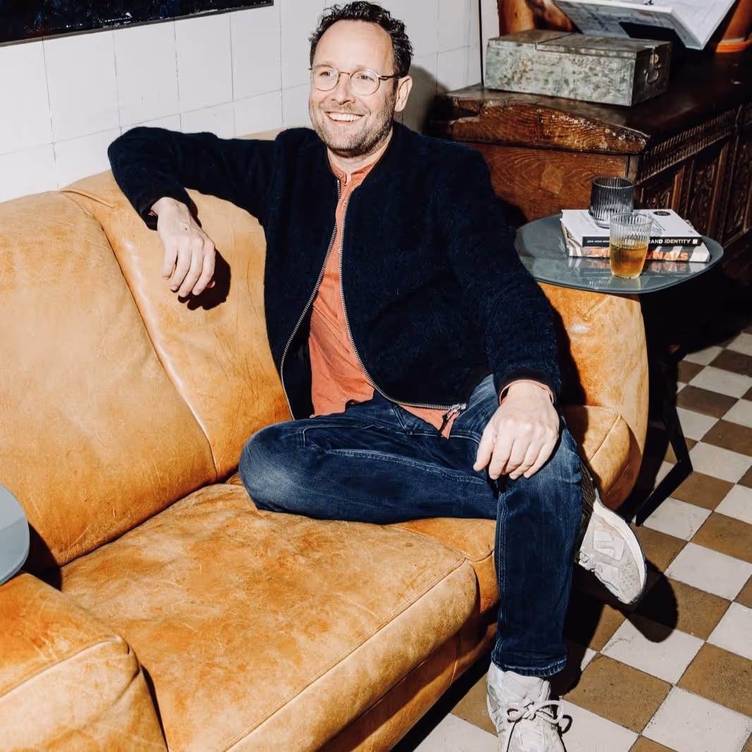 Man met bril en donkere jas zit ontspannen op een bruine leren bank in een kamer met tegelvloer.