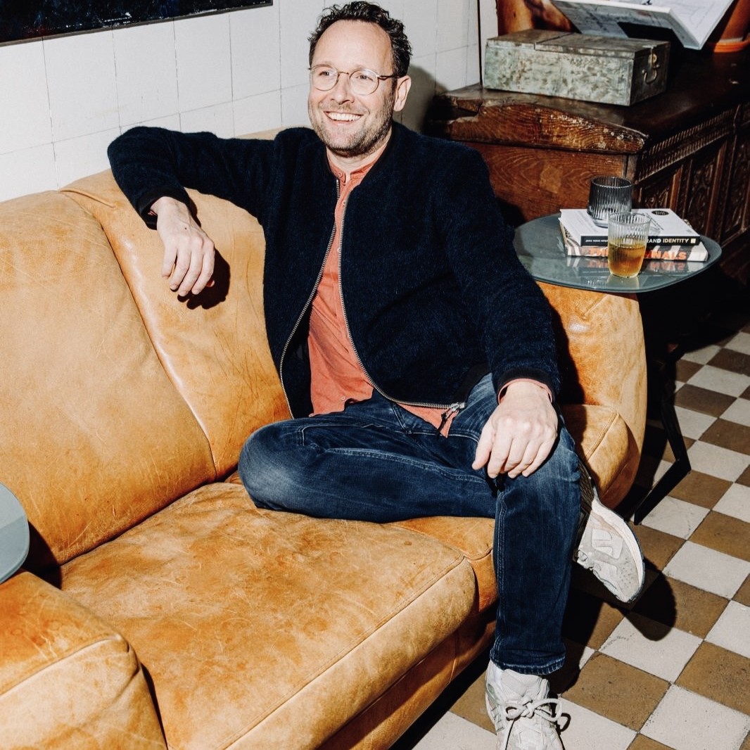 Man met bril en donkere jas zit ontspannen op een bruine leren bank in een kamer met tegelvloer.