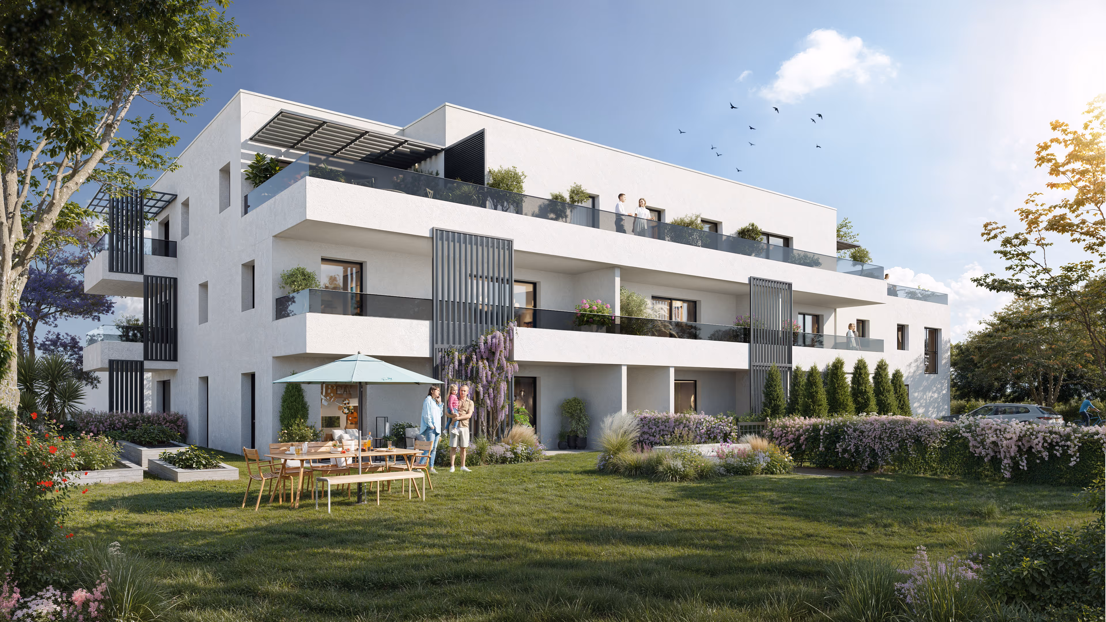 Modern white three-story apartment building with balconies, a lawn with outdoor dining furniture under an umbrella, and a family standing nearby.