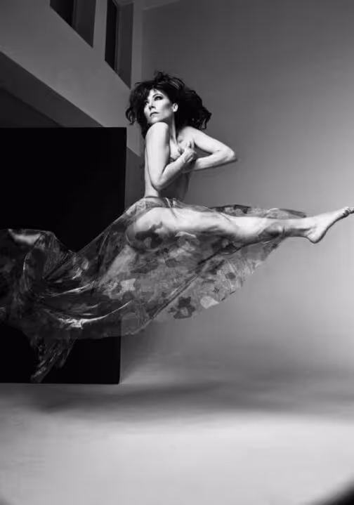 Black and white photo of a female dancer mid-air in a dramatic side leap wearing a flowing skirt.