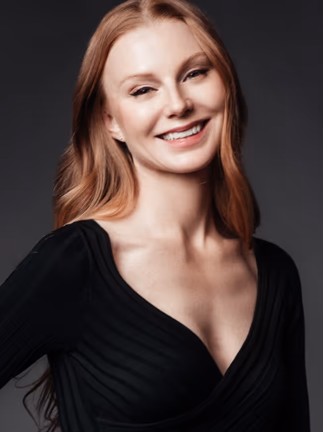 Smiling woman with long red hair wearing a black V-neck top against a dark background.