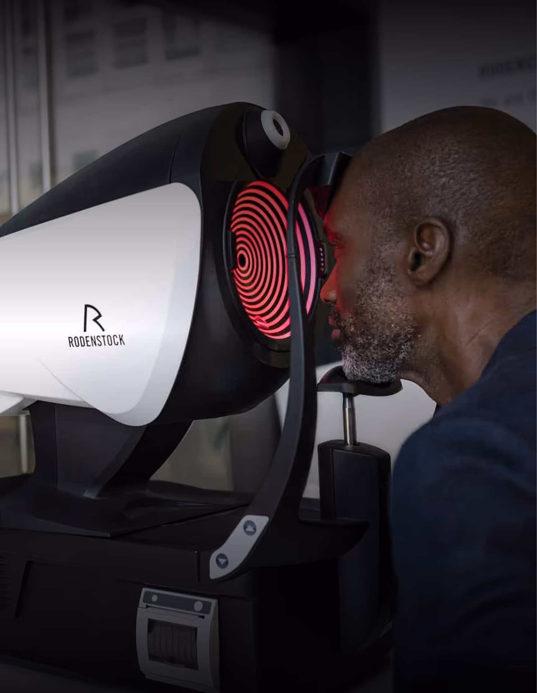 Man undergoing an eye examination using a Rodenstock optical diagnostic machine with red concentric circles visible inside the device.