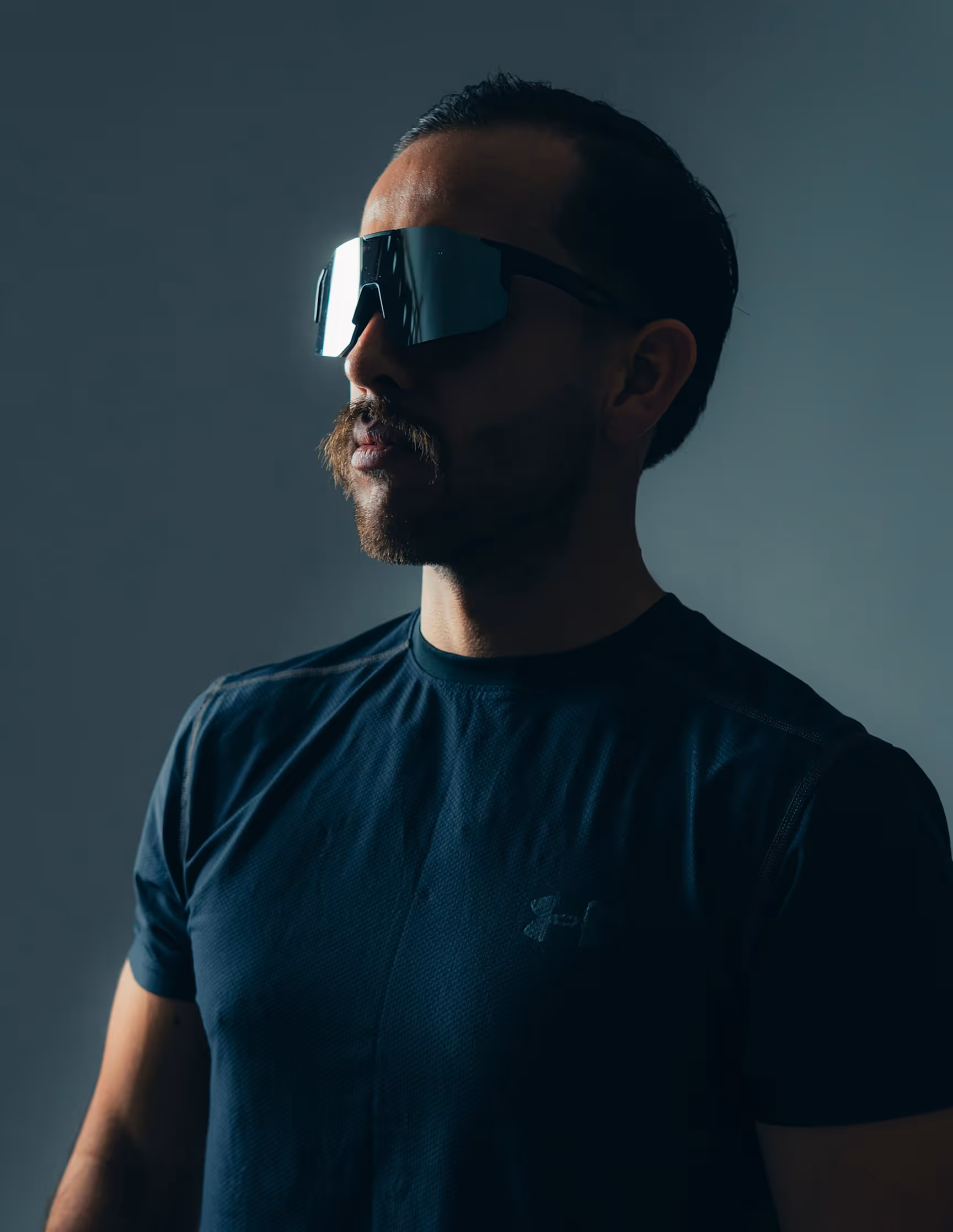 Man with a beard wearing large reflective sports sunglasses and a dark athletic shirt against a muted gray background.