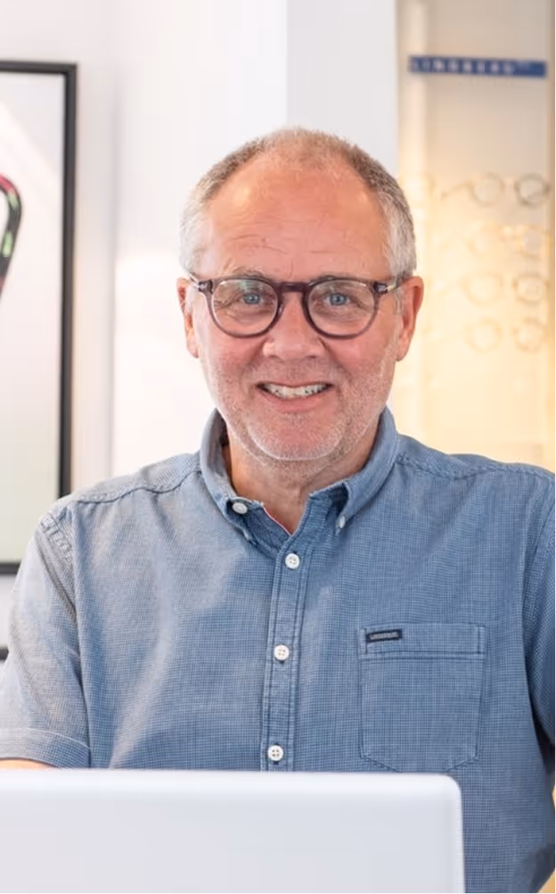 Smiling middle-aged man with glasses wearing a blue button-up shirt, sitting in front of a white laptop.