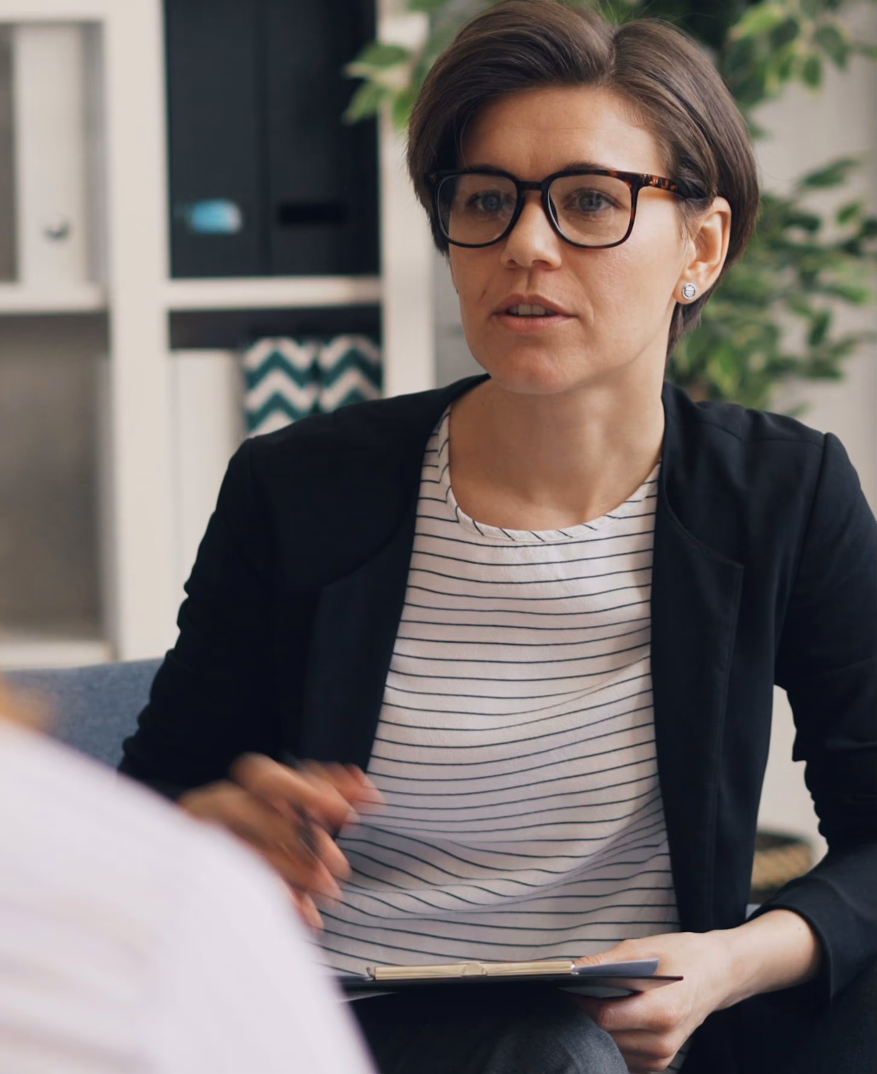 Professional woman with short hair and glasses speaking while holding a clipboard in an office setting.