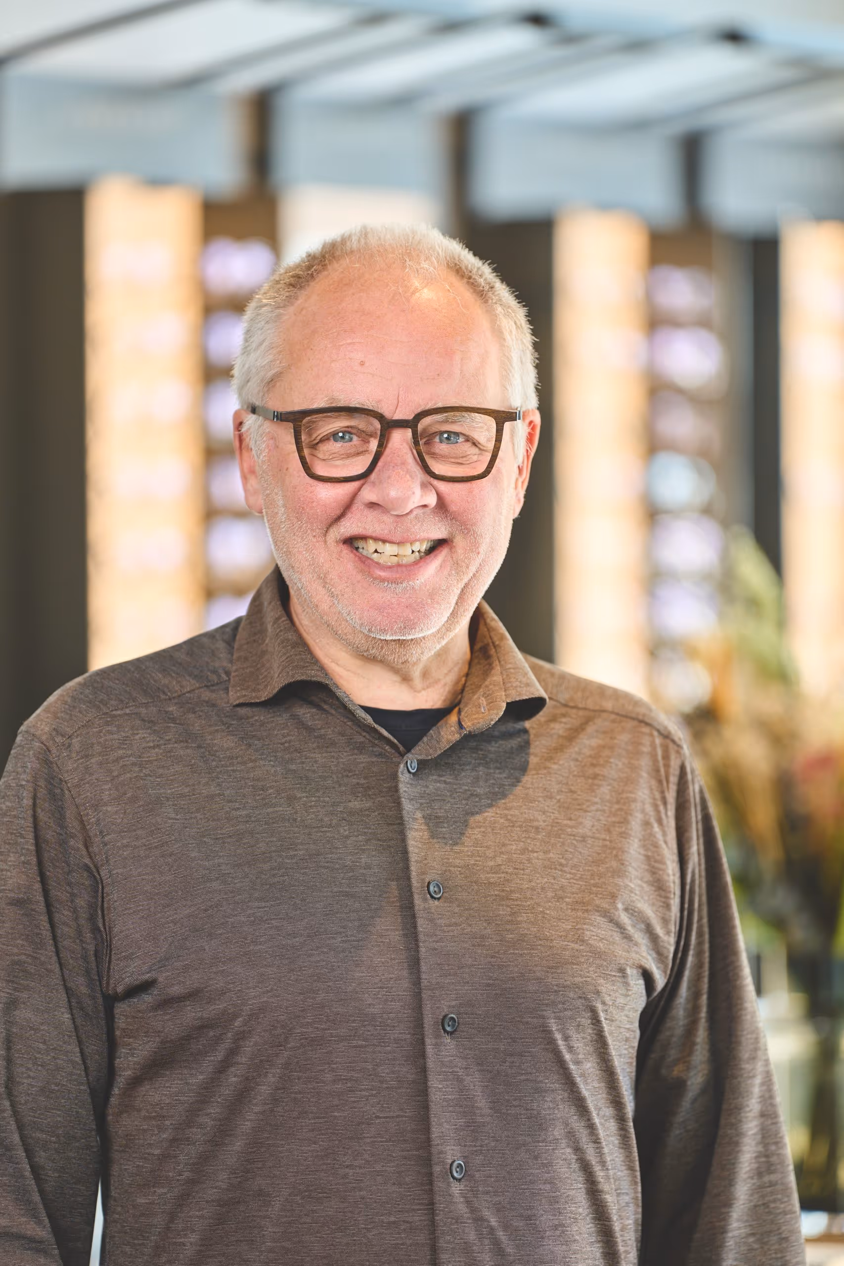 Smiling elderly man with glasses wearing a brown button-up shirt in an indoor setting.